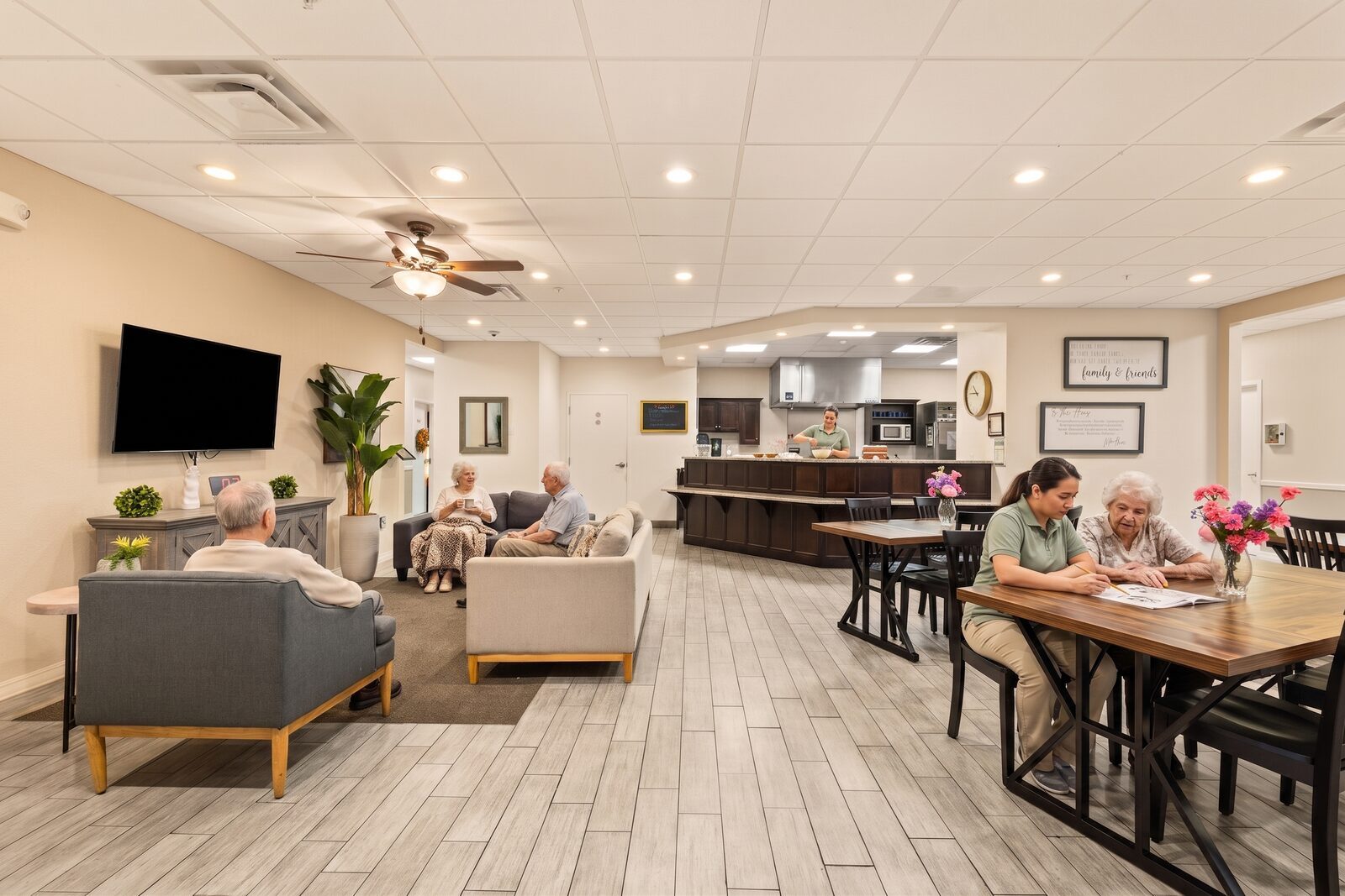 Residents relaxing in the household living room while a caregiver bakes in the open kitchen and another helps with a crossword at the dining table